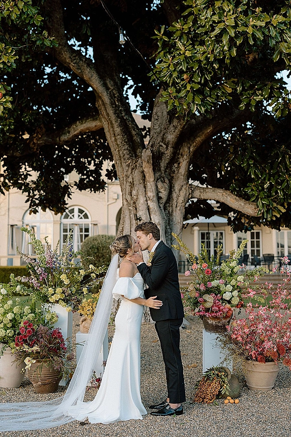 Wedding kiss portrait of bride and groom kissing under a large tree, her veil trailing and string lights glowing in a garden courtyard