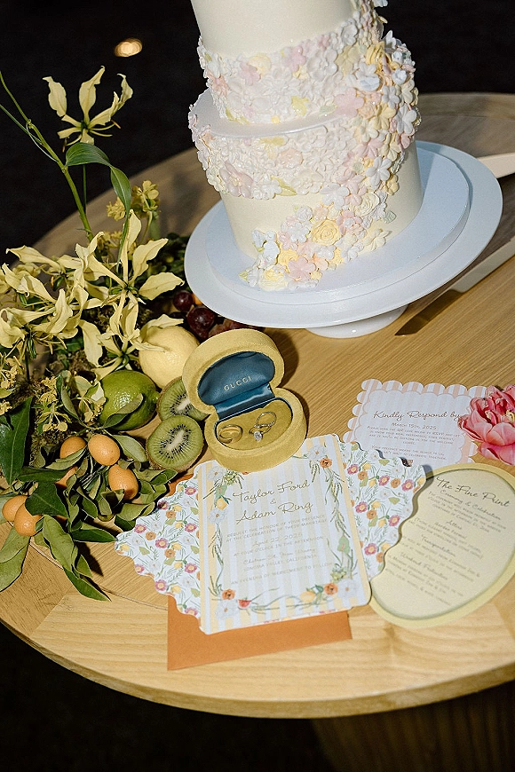 Wedding flat lay featuring a wedding invitation suite with rings, scalloped stationery, fruit, and buttercream cake on a dark wood table