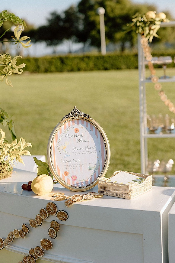 Cocktail menu sign in an ornate gold frame on a white outdoor bar counter with citrus garland, florals, and glassware by the lawn trees