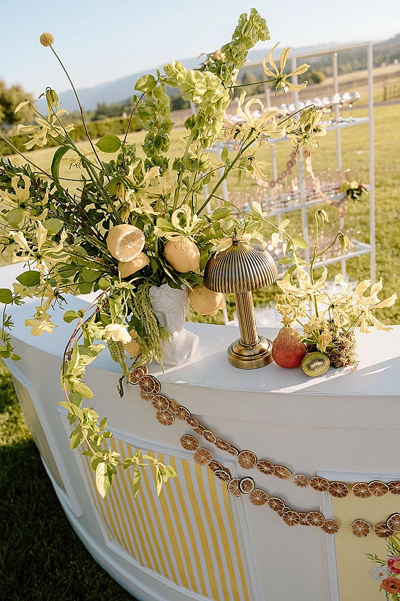 Wedding bar decor with outdoor wedding bar setup featuring citrus florals, lemons, pear and kiwi on a striped counter with brass lamp on lawn backdrop