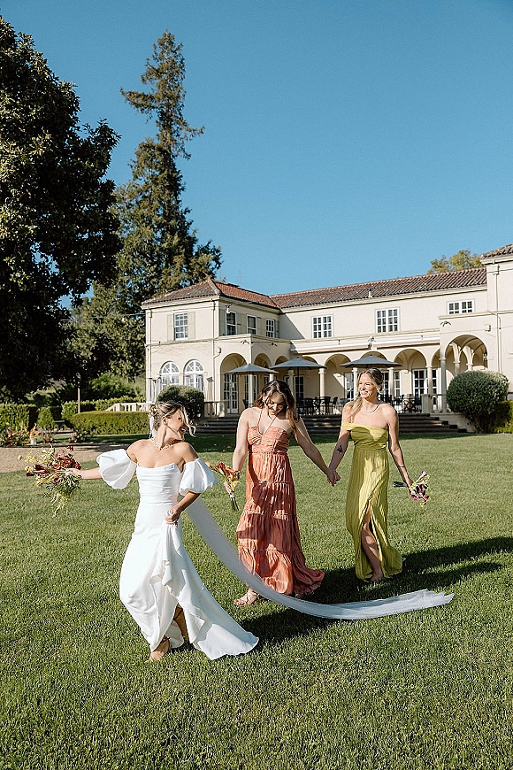Bridesmaid photo of bride with bridesmaids holding hands, carrying bouquets and a long veil, walking on a lawn by a villa under string lights