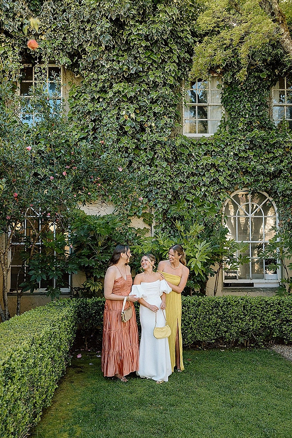 Bridesmaid group photo of the bride with bridesmaids in mismatched dresses, posing by an ivy-covered wall with garden shrubs and windows behind