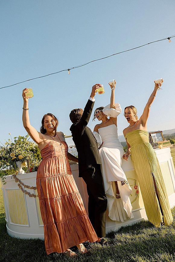 Wedding toast moment as bride and groom cheer with champagne coupes and cocktails at an outdoor bar under string lights, hills beyond
