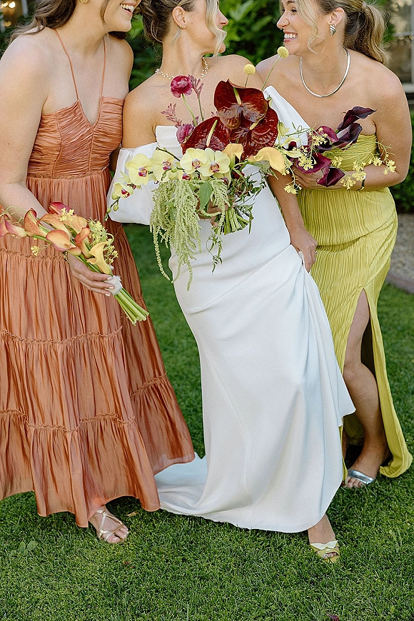 Bride with bridesmaids holding bouquets of orchids, calla lilies and anthurium, smiling on an outdoor lawn by a stone pathway