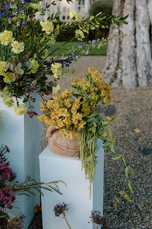 Ceremony floral arrangement in a terra cotta vase on a white plinth, with draping greenery and blue, yellow, purple blooms outdoors