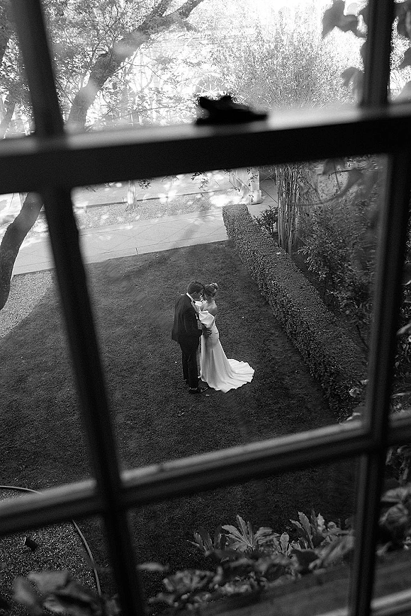 Wedding couple portrait in a black and white wedding photo, bride and groom kiss seen through window panes with garden lawn beyond
