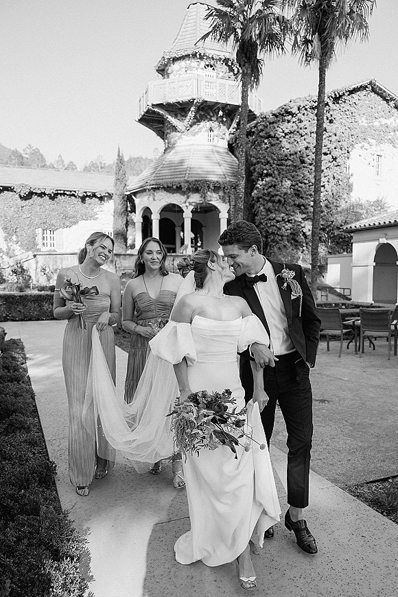 Wedding party portrait of bride and groom walking with bridesmaids behind, bouquet and veil in an ivy-covered historic courtyard walkway