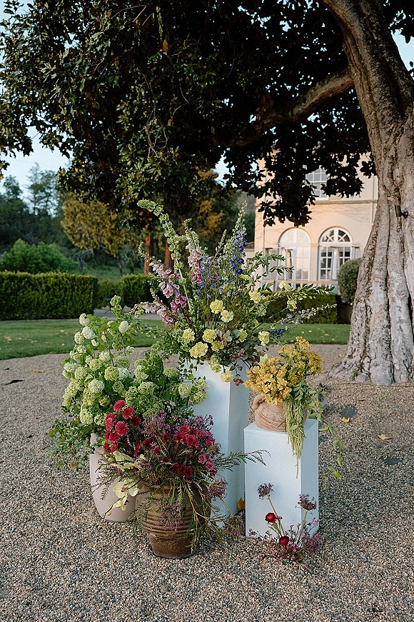 Wedding floral arrangements in ceramic urns on white pedestals with lush greenery lining a gravel courtyard under a large tree
