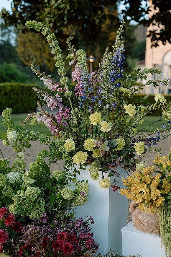 Wedding floral arrangement of blue, purple, and yellow garden blooms with greenery in a ceramic urn on a pedestal, set on a lawn near hedges and a stone wall