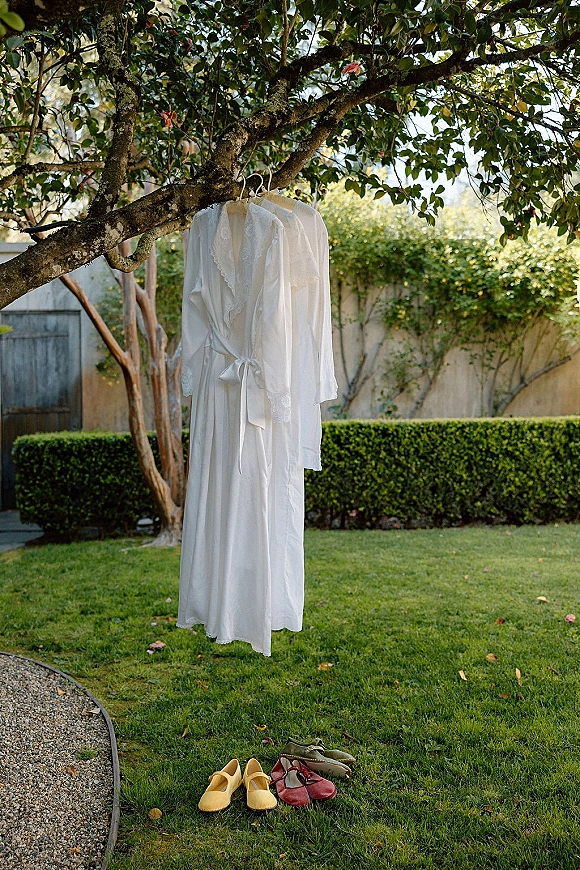 Bridal robe with lace trim hanging from tree branches in a garden, with shoes below and a stucco wall and wooden gate behind