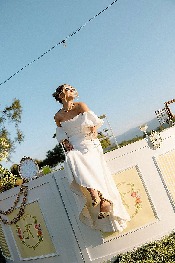 Bridal portrait of a bride in sunglasses in an off the shoulder wedding dress holding a cocktail at an outdoor bar with mountains behind