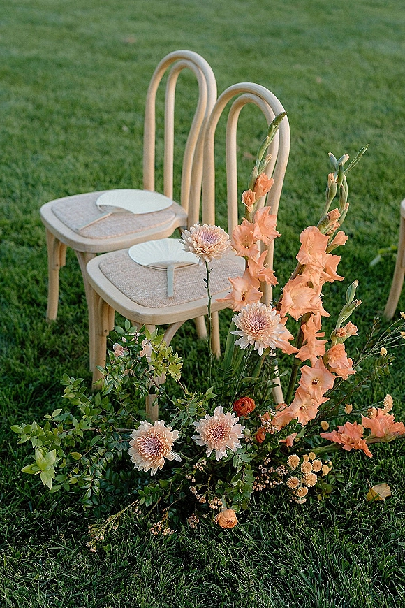 Ceremony chair decor with rattan wedding chairs and handheld fans, beside a peach gladiolus and blush chrysanthemum floral cluster on grass lawn