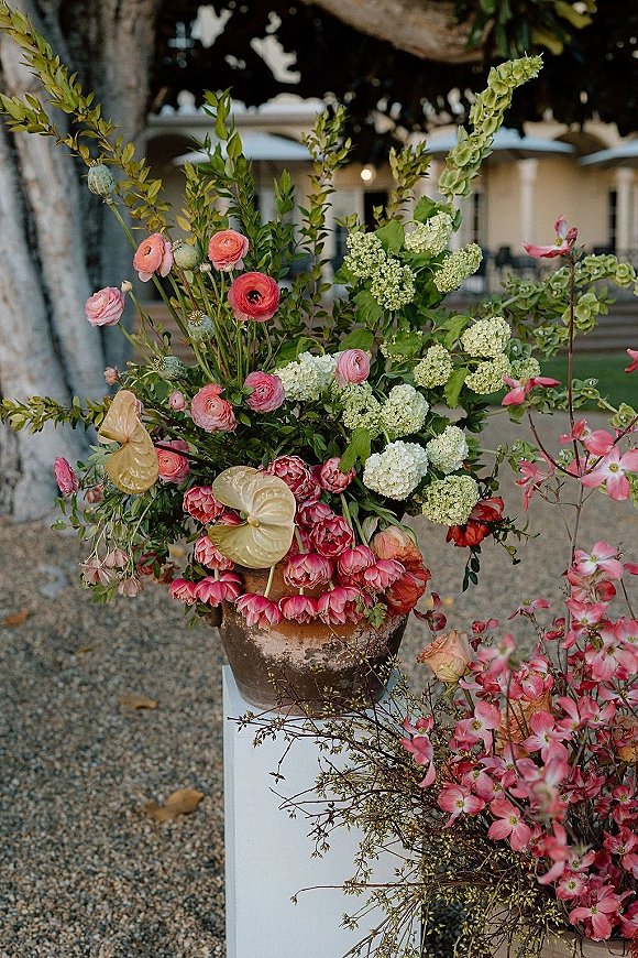 Wedding floral arrangement in a terracotta urn on a pedestal, with pink ranunculus and white hydrangea beside a porch entrance