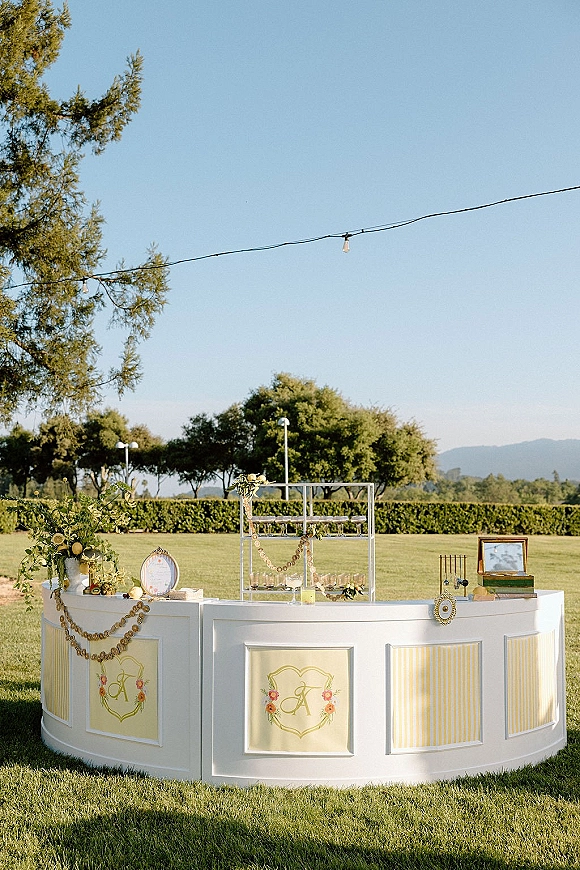 Wedding bar setup with a curved outdoor wedding bar, greenery garland, citrus decor, glassware shelves, and string lights on a lawn with mountains