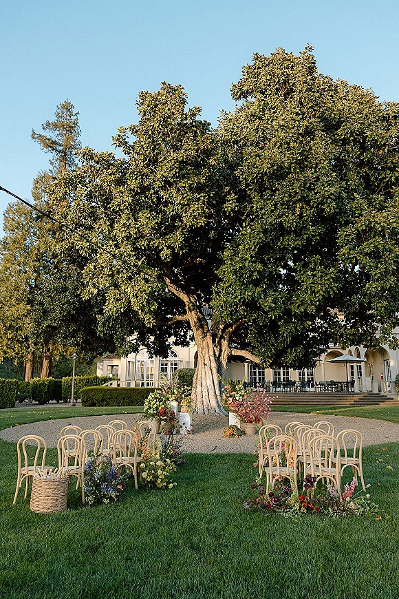 Outdoor ceremony setup with garden wedding ceremony seating in the round, curved gravel aisle lined with wildflower florals beneath a large tree