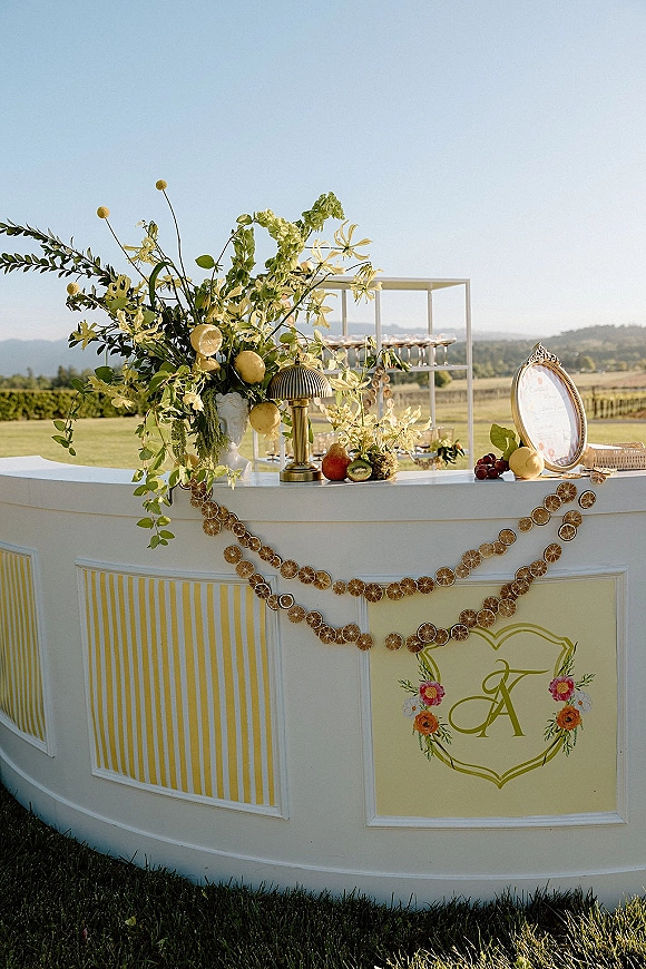 Wedding bar setup with a white outdoor wedding bar, dried orange garland, citrus and florals, plus menu sign on a vineyard lawn backdrop