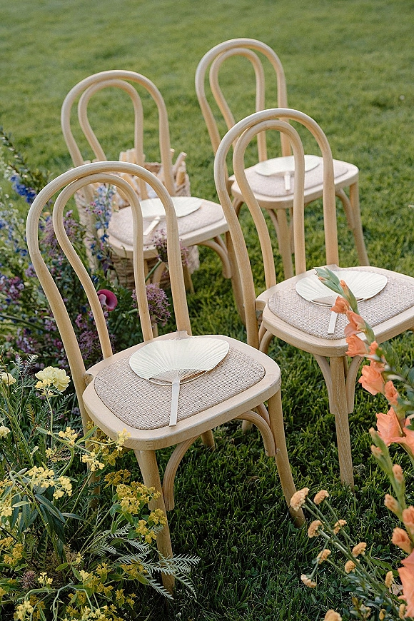 Ceremony seating with outdoor ceremony chairs, bentwood and woven seats, handheld fans, and florals with greenery on a grass lawn