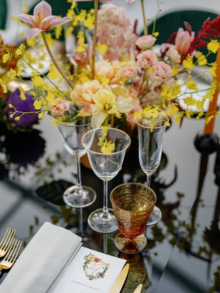 Reception tablescape with a wedding table centerpiece of pastel flowers and yellow orchids, amber goblet, gold flatware, on dark glass table with green velvet chair