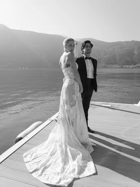 Couple portrait in black and white, bride in strapless lace gown with long train and groom in tuxedo on a dock by lake and mountains
