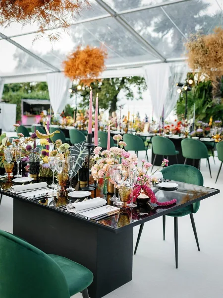 Reception tablescape with modern wedding table setting on a black table, green velvet chairs, floral centerpieces, and pink taper candles under a clear tent
