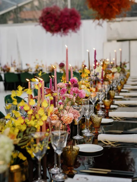 Reception tablescape with wedding table centerpiece of colorful florals, taper candles, and amber goblets before a white draped backdrop