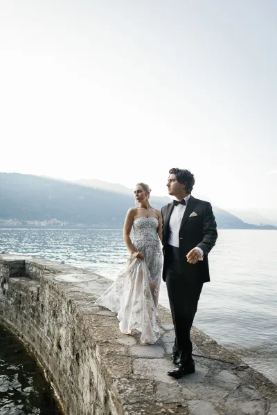 Couple portrait of bride and groom walking on a stone walkway by a mountain lake, bride holding her dress train in a beaded gown