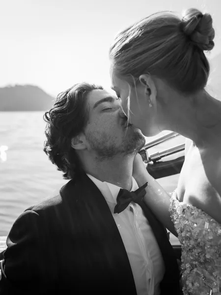 Wedding kiss as bride in a strapless beaded gown and groom in tuxedo embrace on a boat, with mountain lake and sky behind