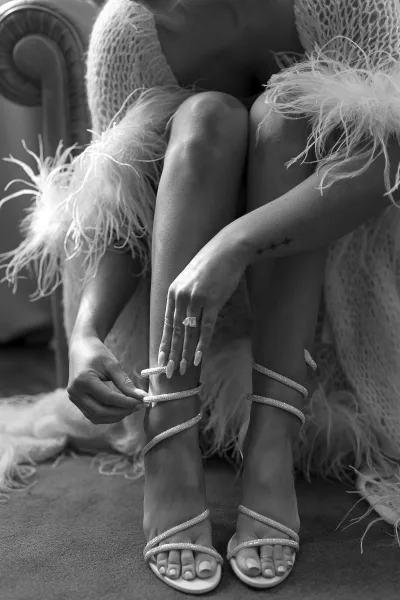 Bridal shoes, rhinestone strappy heels on the bride’s feet as she puts them on, showing engagement ring and manicure in a bedroom setting