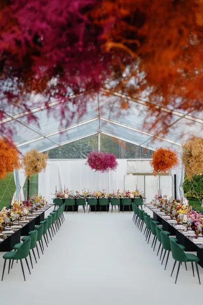 Reception tablescape with a long banquet table wedding setup, green velvet chairs, amber goblets, taper candles and hanging florals in a clear tent on grass