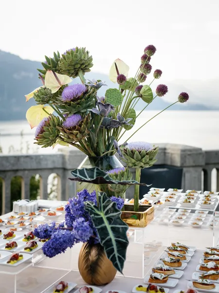 Cocktail reception table with wedding cocktail hour appetizers on white plates and acrylic risers, tall gold vase centerpiece on a lakeside terrace