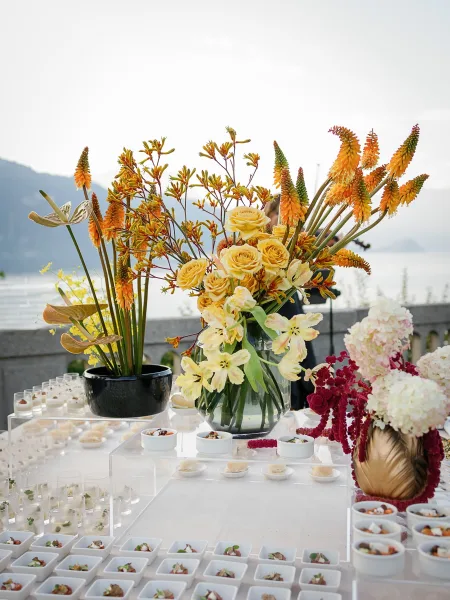 Wedding cocktail table with white acrylic risers, shot glass appetizers, and a tall yellow rose floral centerpiece on a terrace by the lake and mountains