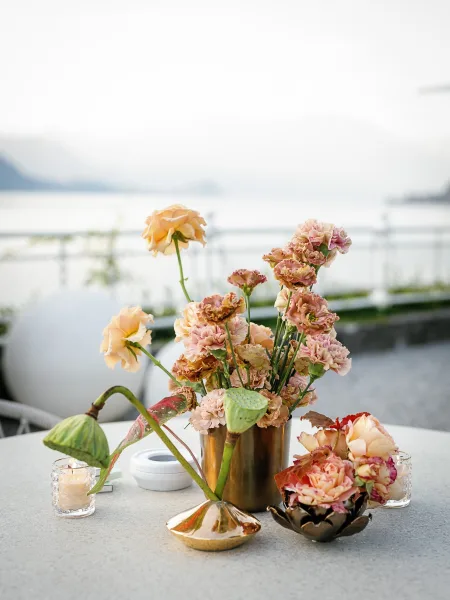 Wedding centerpiece in a gold vase with roses, carnations, lotus pods, and glowing glass votive candles on a terrace by the water