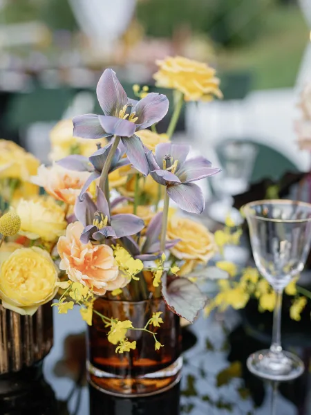 Wedding centerpiece with purple tulips and yellow roses in an amber glass vase, set among stemware on an outdoor lawn banquet table
