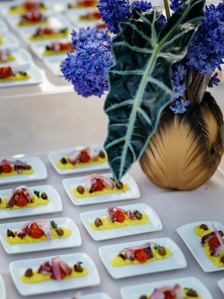Wedding appetizer display featuring plated octopus bites with yellow puree, olives, and herb garnish beside a purple floral centerpiece in gold vase