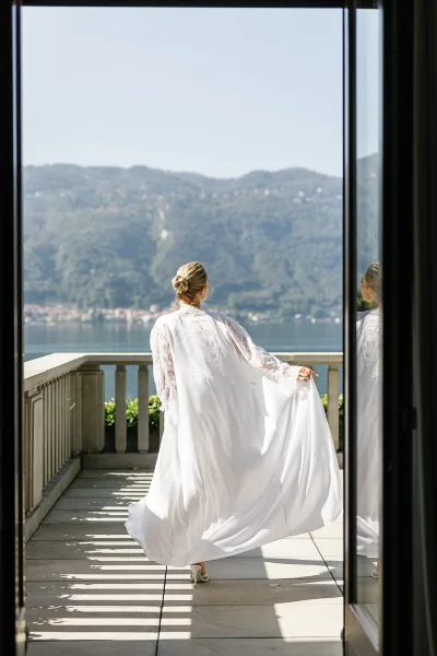Bridal portrait of a bride from behind in a lace sleeve wedding dress, holding her train on a balcony terrace overlooking a mountain lake
