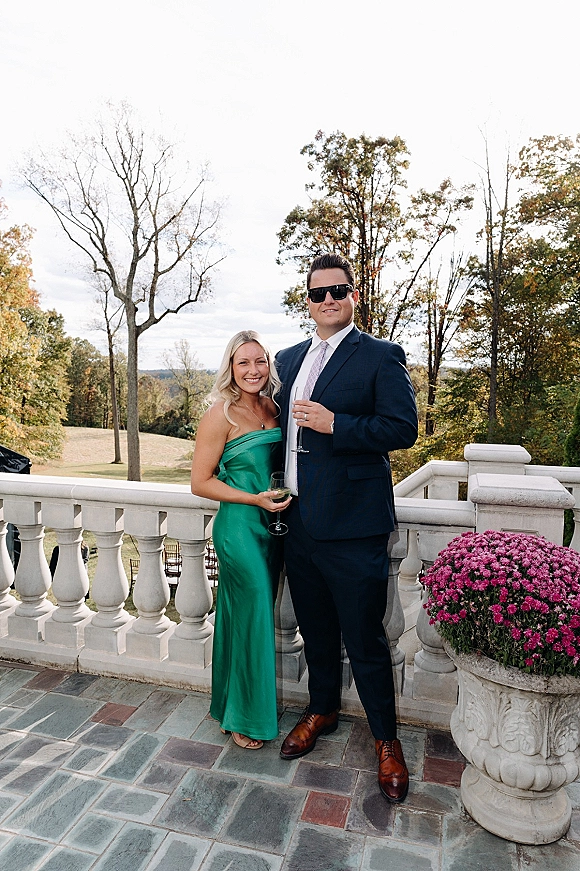 Wedding guest portrait of a couple on an outdoor terrace, one in an emerald green satin dress and the other in a navy suit, holding drinks