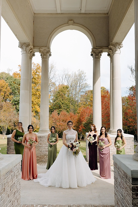 Bride with bridesmaids in a bridal party portrait, holding bouquets beside a white-columned portico with autumn trees behind
