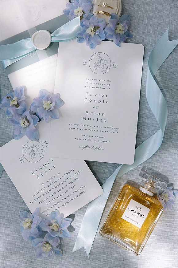 Invitation suite wedding invitation flat lay with blue ribbon and wax seal, rings and perfume arranged on light blue linen with blue flowers
