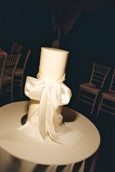 Wedding cake, a white wedding cake wrapped with a satin ribbon, set on a round table with chiavari chairs receding into a dark background.