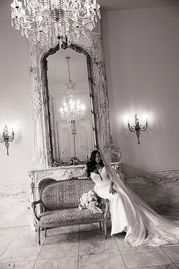 Bridal portrait in a strapless wedding dress, bride seated on an antique sofa with long veil and bouquet in a marble hallway with chandelier