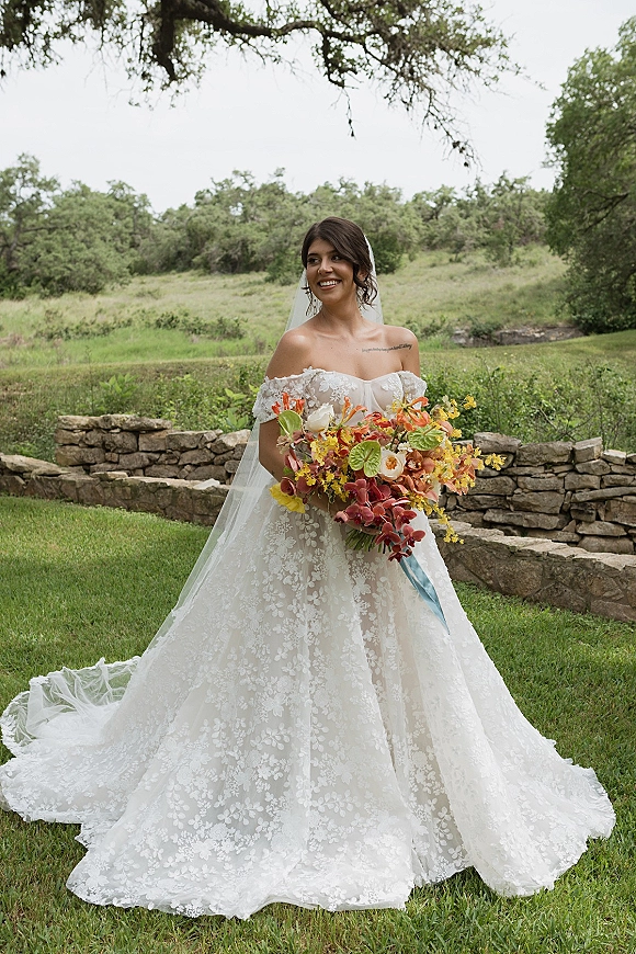 Bridal portrait of a bride in an off the shoulder lace wedding dress holding a colorful bouquet with blue ribbon by a stone wall lawn