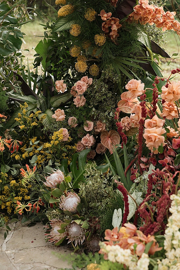 Wedding floral installation ceremony floral backdrop with peach orchids and roses, palm leaves, and draped fabric on a garden lawn