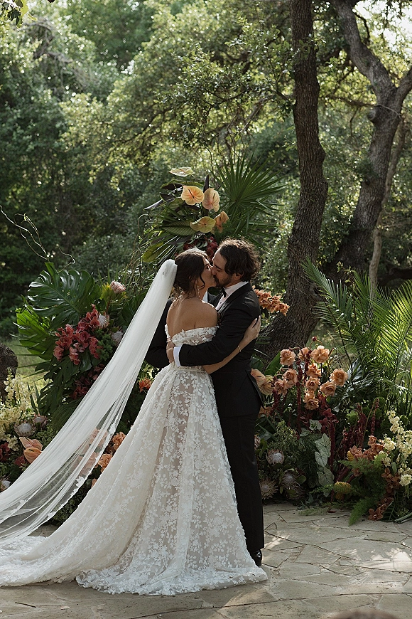 Wedding kiss portrait of bride and groom kiss, her lace off-shoulder gown and cathedral veil beside tropical florals on a stone patio in trees