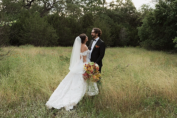 Couple portrait of bride and groom standing in a wildflower meadow, gazing at each other as she holds a rust bouquet and veil flows