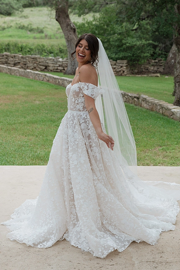 Bridal portrait in an off the shoulder wedding dress with lace sleeves, long veil and flowing train on a green garden lawn by trees
