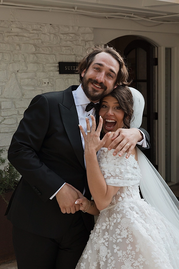 Wedding couple portrait with bride showing engagement ring as she laughs with groom in black tuxedo by a stone wall doorway outside