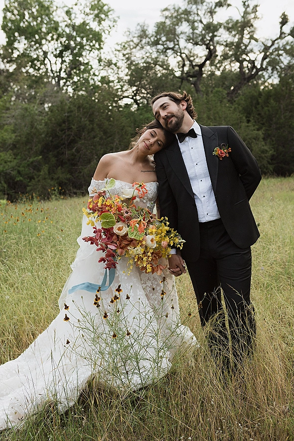 Couple portrait of bride and groom holding hands, her off-shoulder lace dress and colorful bouquet in a wildflower meadow under overcast sky