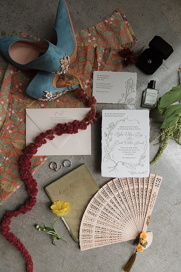 Invitation suite wedding invitation flat lay with RSVP card, envelopes, wax seal, rings, blue shoes, perfume, and red floral garland on concrete surface
