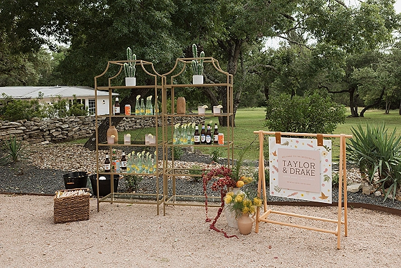 Wedding welcome sign on a wooden stand beside a custom wedding welcome sign bar shelf with bottles, candles, cactus, and flowers on a gravel patio