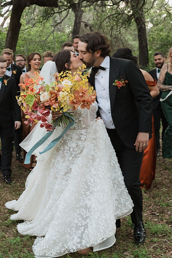 Wedding kiss as the bride and groom kiss, bride holding a bouquet with blue ribbon while bridesmaids and groomsmen stand on grass by trees